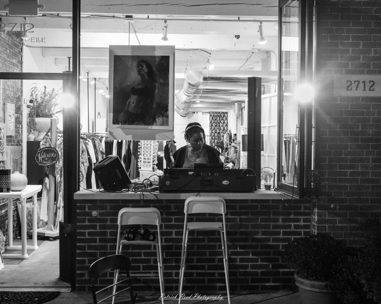 A DJ wearing headphones works behind an open storefront window at Eastern Market After Dark, photographed in black and white.