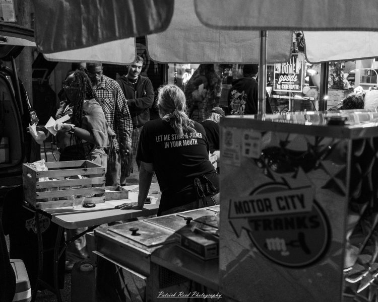 Customers gather at the Motor City Franks food stand beneath an umbrella at Eastern Market After Dark, photographed in black and white.