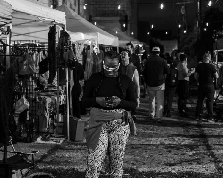 A person looks down at a phone between illuminated vendor stalls at Eastern Market After Dark, photographed in black and white.