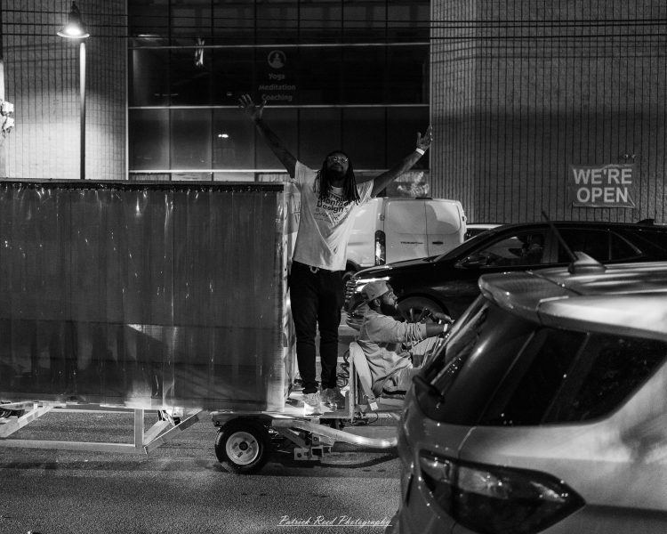 A person raises both arms beside a mobility scooter and parked vehicles at Eastern Market After Dark, photographed in black and white.