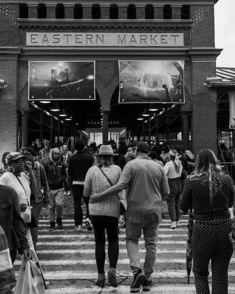 A couple walks toward the Eastern Market entrance beneath two large photographs, surrounded by visitors in a black-and-white street scene.