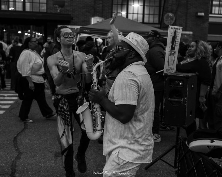 A saxophonist in a light hat performs beside a dancer in the crowd at Eastern Market After Dark, photographed in black and white.