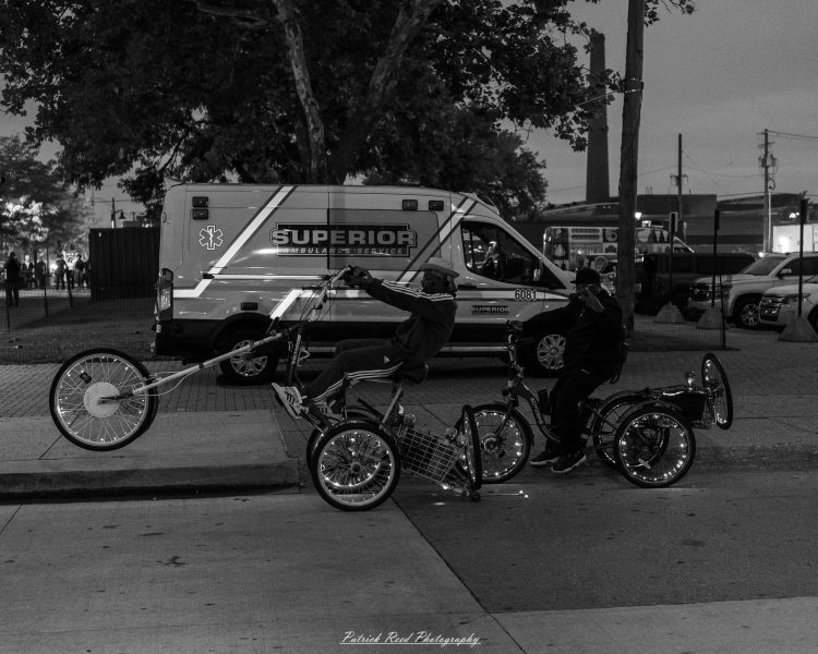 Two riders on custom cycles pass a parked Superior ambulance at Eastern Market After Dark, photographed in black and white.