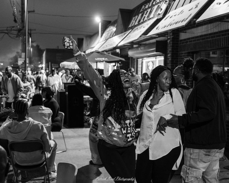 Two people pose together as one raises a phone above the crowd at Eastern Market After Dark, photographed in black and white.