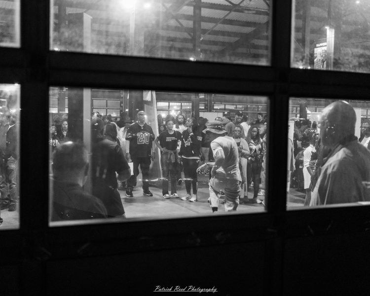 Saxophonist in a light hat performs at an after-dark street music set with a watching crowd.