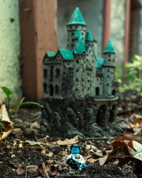 Miniature medieval castle model with turrets and blue roofs beside a LEGO-style figure outdoors.