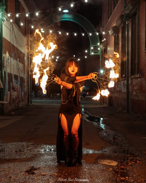 A fire dancer stands in the center of a dark brick alley at night, extending both arms while spinning flaming fans. Orange firelight illuminates her face and black outfit, while overhead string lights and reflections on the wet pavement frame the scene.