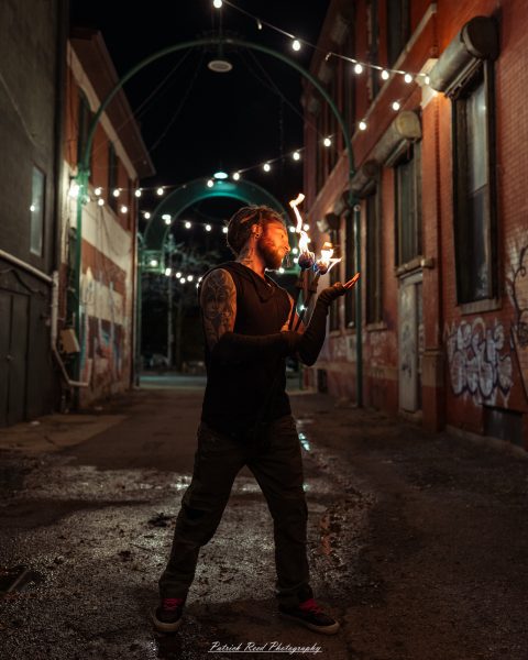 A tattooed, bearded fire performer stands in a narrow brick alley at night, holding a flaming prop near his face with one hand extended beneath it. Warm firelight illuminates his face and black clothing, while overhead string lights, graffiti-covered walls, and wet pavement fill the background.