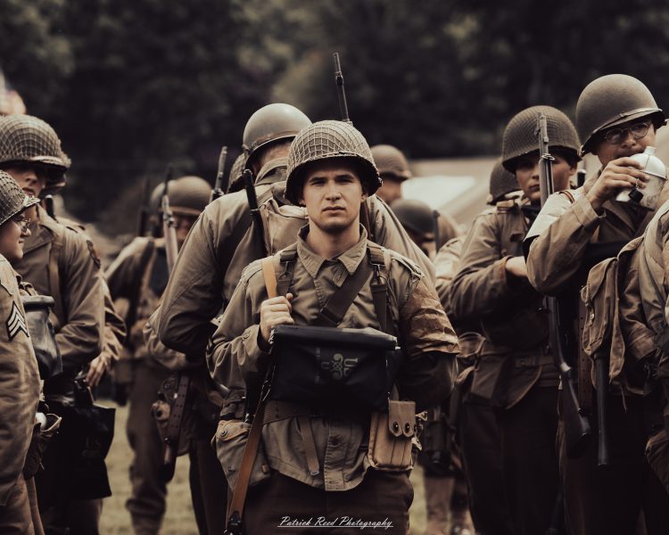 A young World War II reenactor in a netted helmet looks toward the camera amid a group of uniformed participants.
